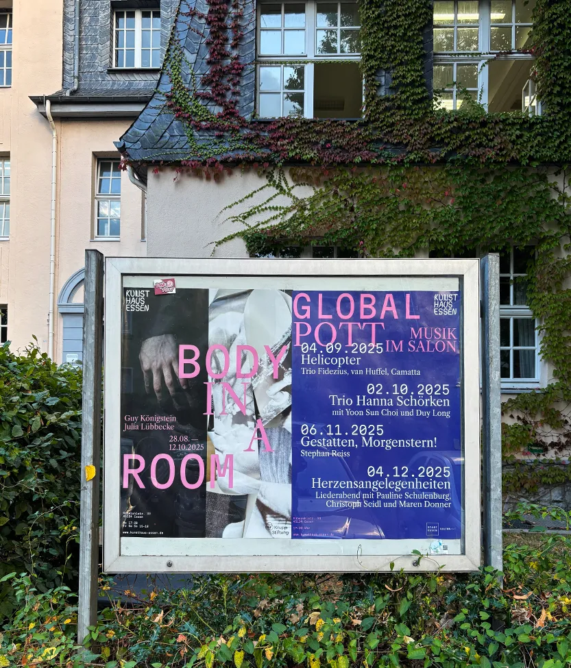 Eine rote Rennradlehne lehnt an einer Betonwand mit drei bunten Veranstaltungspostern für Kunsthaus Essen.