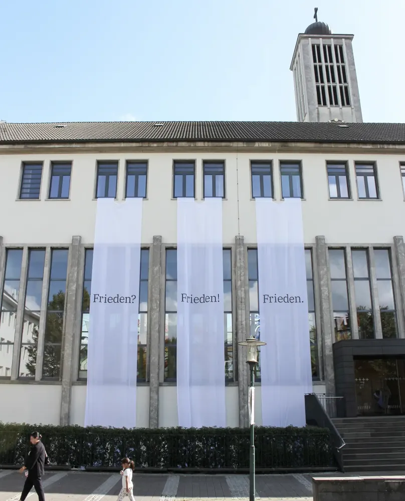 Eine rote Rennradlehne lehnt an einer Betonwand mit drei bunten Veranstaltungspostern für Kunsthaus Essen.