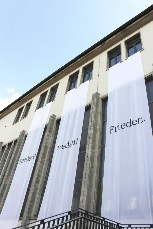 Weiße Banner mit den Worten ‚Frieden?‘, ‚Frieden!‘ und ‚Frieden.‘ hängen an der Fassade eines Gebäudes mit mehreren Fenstern.
